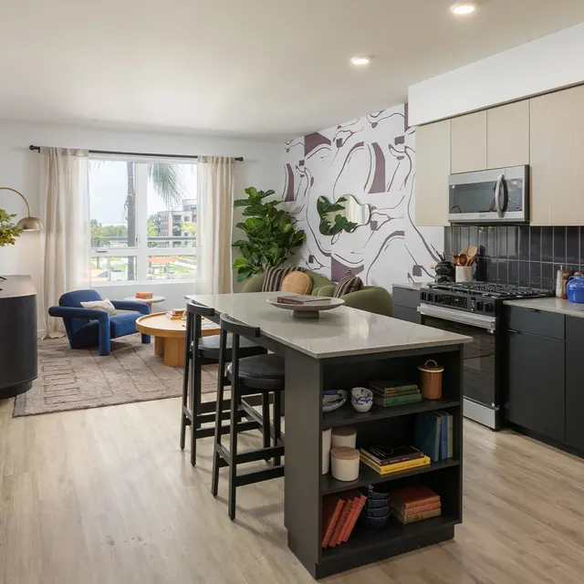 A modern kitchen and living room space featuring an island countertop, black and wooden cabinetry, a dining table, and a decorative wall with plants.