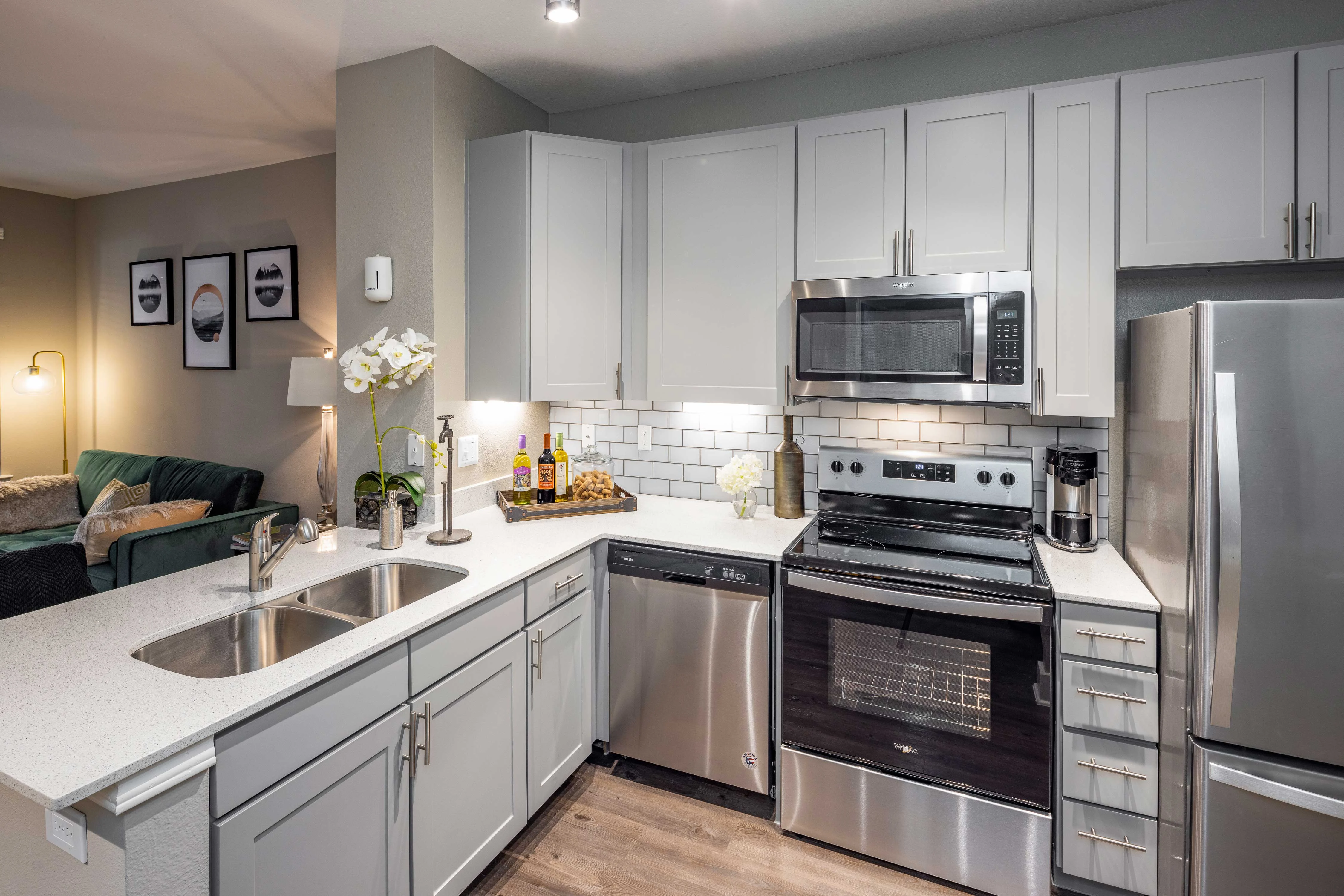 Modern Kitchen Interior A contemporary kitchen with light gray cabinets, stainless steel appliances, and a white countertop. The kitchen features a double sink, an oven, a microwave, and a dishwasher, arranged in an L-shape. Decorative elements include a wine bottle display and a small vase with white flowers.