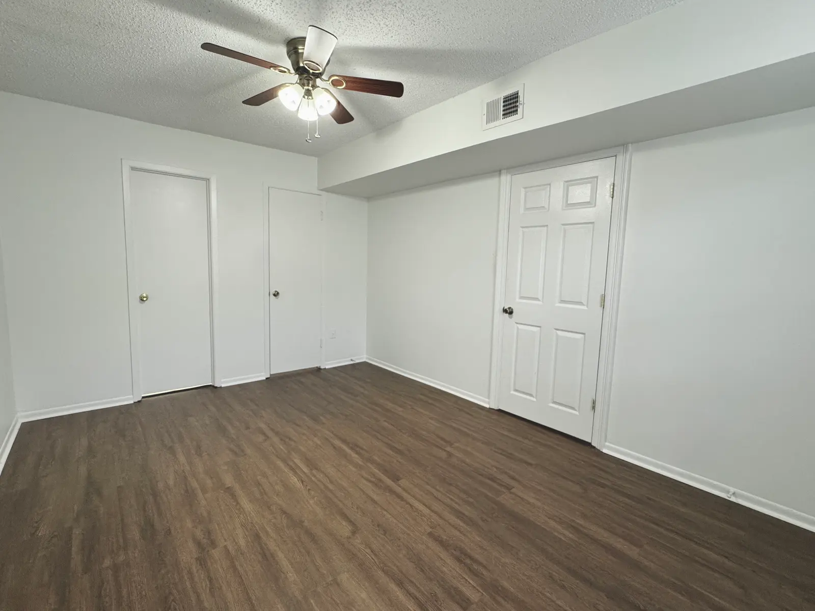An empty room featuring light wood flooring, two closed doors on the left wall, a door on the right wall, and a ceiling fan with light fixtures in the center of the ceiling.