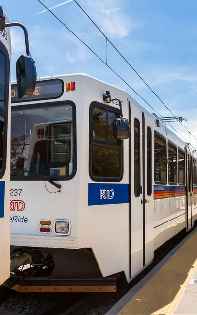 A light rail vehicle at a station with a covered waiting area and a sunny sky in the background.