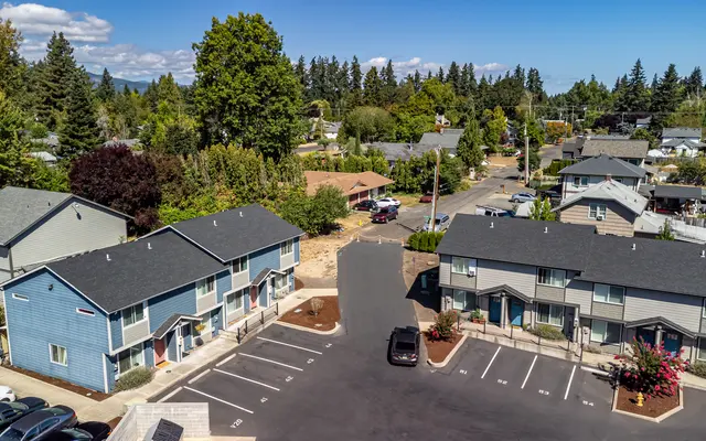 Aerial view of a residential area featuring a housing complex with multiple units, parking spaces, and surrounding greenery. The landscape is dotted with trees and suburban homes under a clear blue sky.