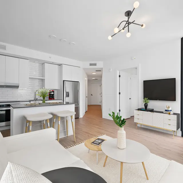A modern apartment interior featuring a light-colored living space with a white couch, a small round coffee table, and bar stools by the kitchen island. The kitchen has sleek cabinets and appliances, and there's a large TV mounted on the wall. Natural light floods the room through large windows.