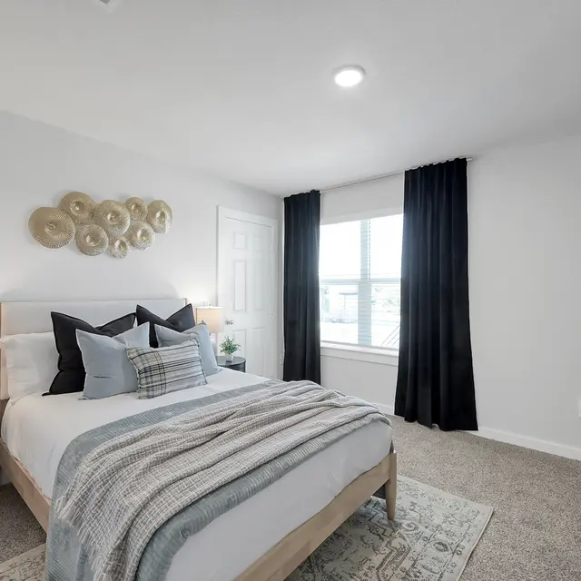 A modern, bright bedroom featuring a bed with gray and white bedding, decorative pillows, a round decorative piece on the wall, and a small nightstand with a lamp. Curtains frame the window, and there is a dresser next to the bed.