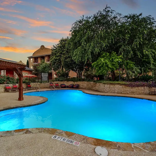 A beautifully lit swimming pool area with glowing blue water, surrounded by greenery and lounge chairs, under a colorful sunset sky.