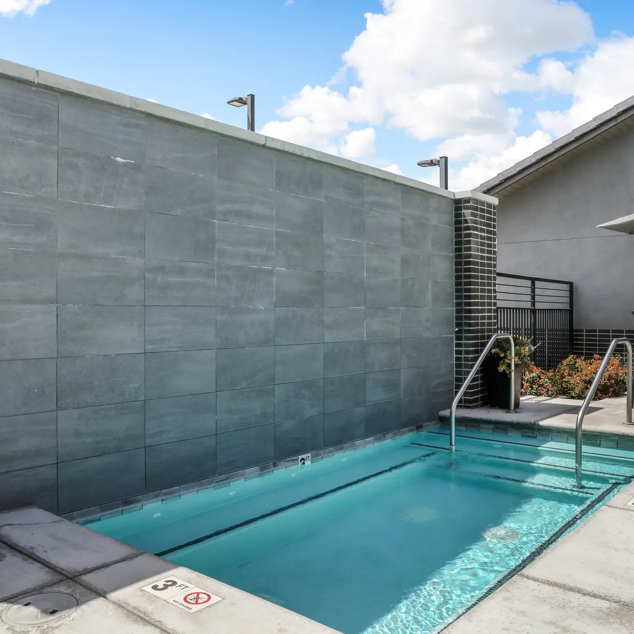 A modern outdoor pool area featuring a small pool with clear water, bordered by gray tiles. There are steps leading into the pool, with a tall wall in the background. The surrounding area has a patio with seating under an umbrella, and decorative plants in planters.