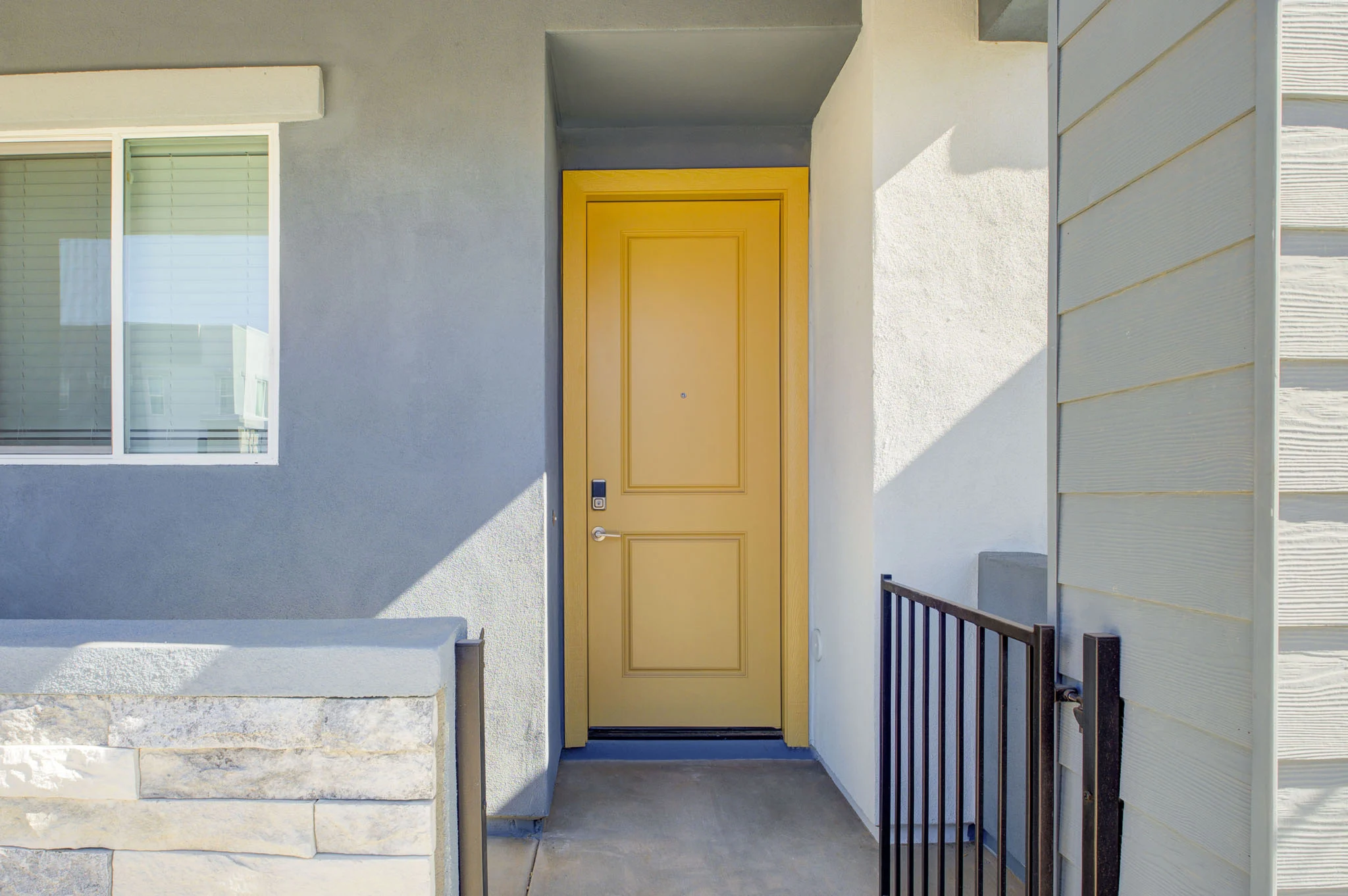 A modern entryway featuring a bright yellow door, gray walls, and a small railing. Next to the door, there's a window that allows natural light to filter in.