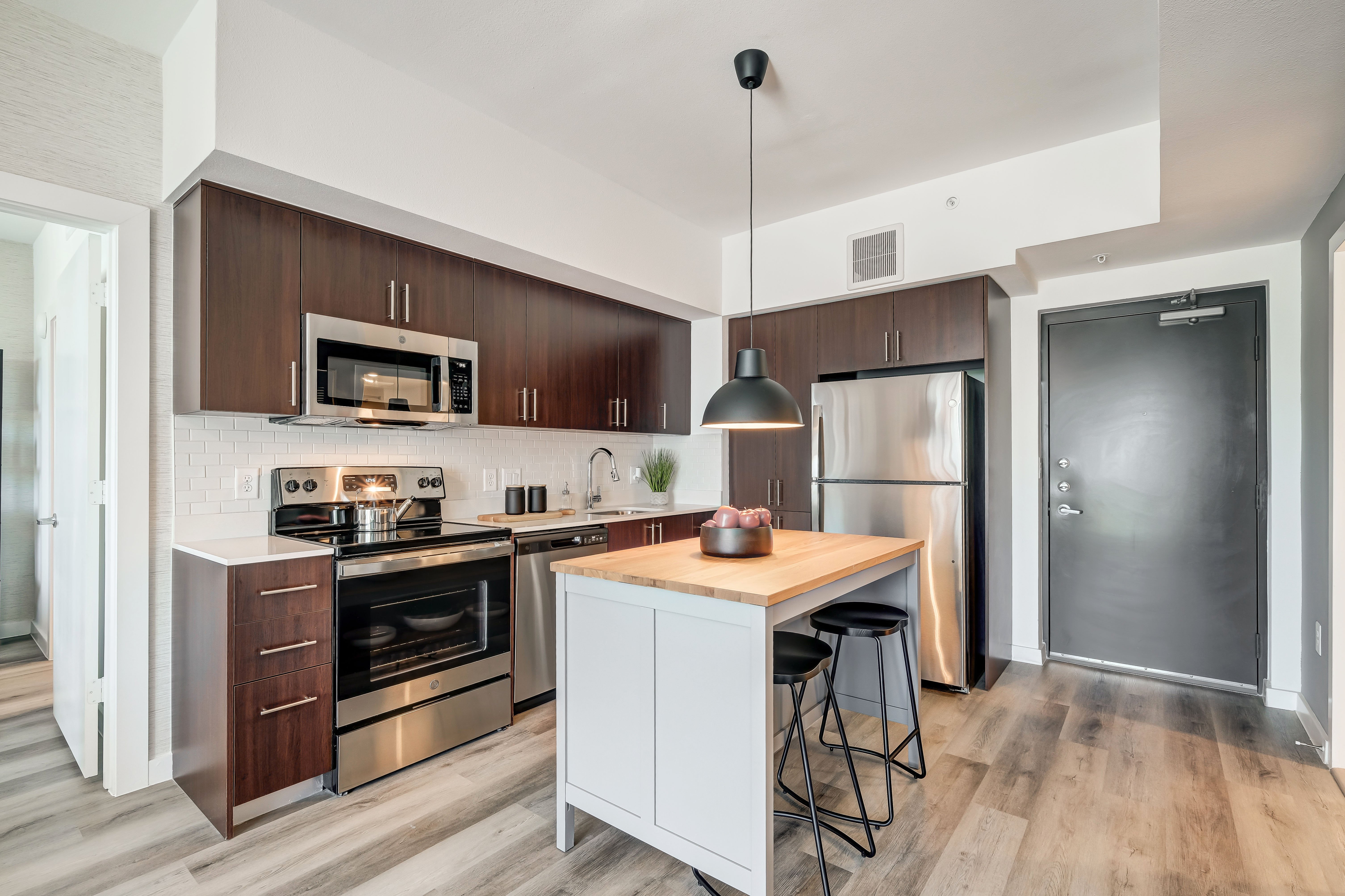 A modern kitchen featuring dark wood cabinets, stainless steel appliances, and a kitchen island with stools. Bright and spacious with a door in the background.