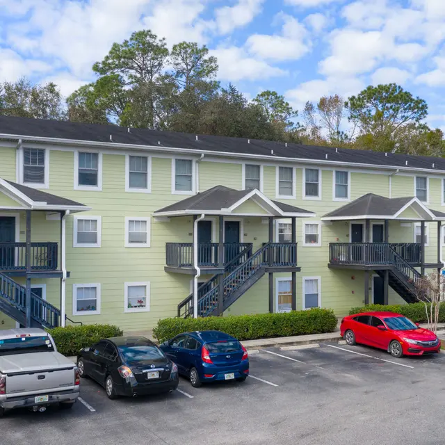A multi-story apartment complex with a light green exterior, featuring balconies and staircases. Several parked cars are visible in front.