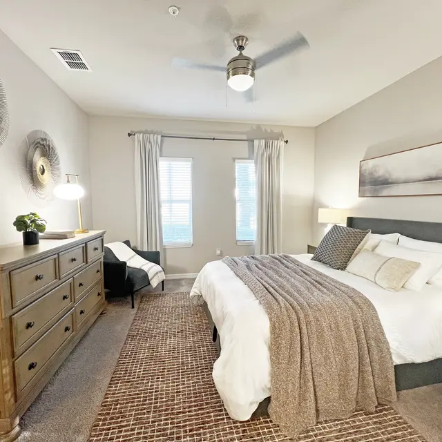 A cozy and modern bedroom featuring a large bed with a grey upholstered headboard, surrounded by soft bedding and a knit throw. There's a wooden dresser with decorative items, a small armchair, and warm lighting. The room has light-colored walls, a ceiling fan, and two windows with curtains.