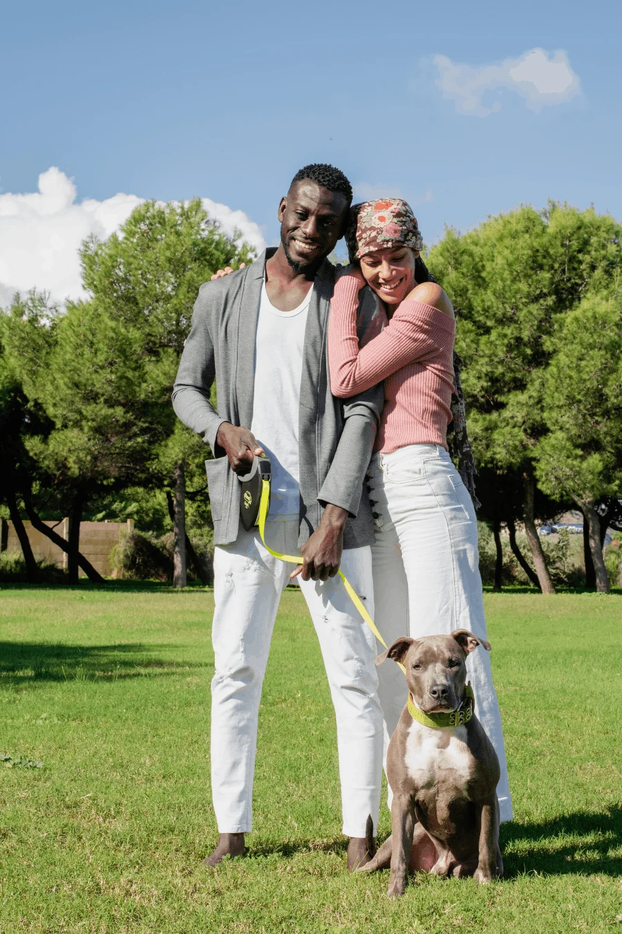A man and a woman posing together in a park with a dog. The man is holding a dog's leash, and they both look joyful. The woman is leaning into the man while smiling. They are surrounded by green grass and trees under a blue sky with clouds.