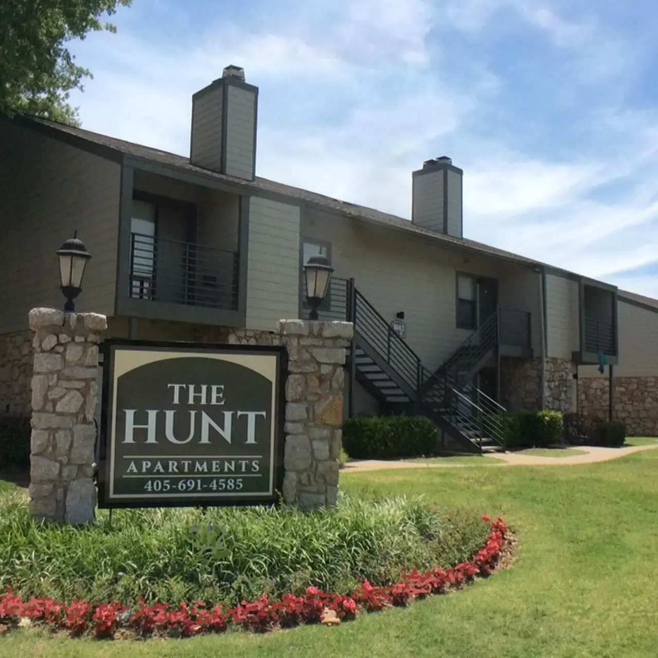 The Hunt Two-story apartment complex with a stone sign in the foreground reading "The Hunt Apartments" and a manicured lawn with flower beds.