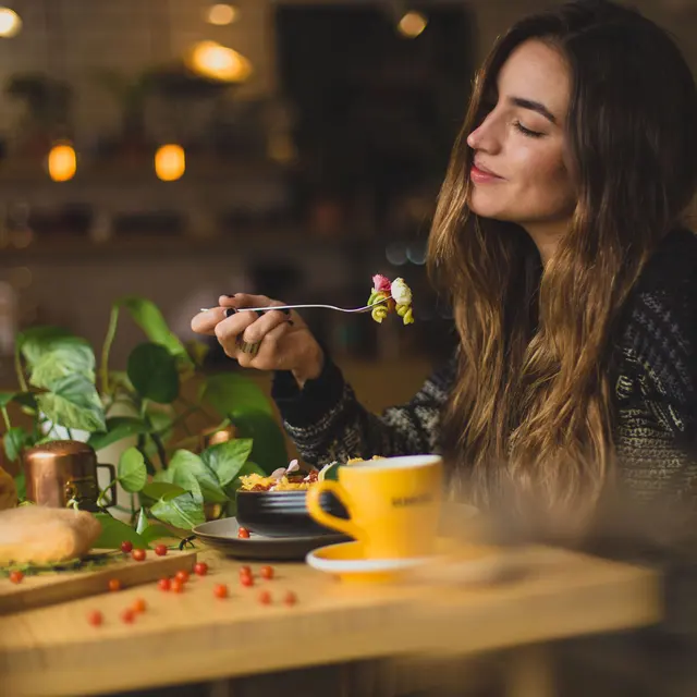High Street Lofts A woman with long hair smiles while enjoying a meal at a cozy, plant-filled café, with a yellow cup in front of her.