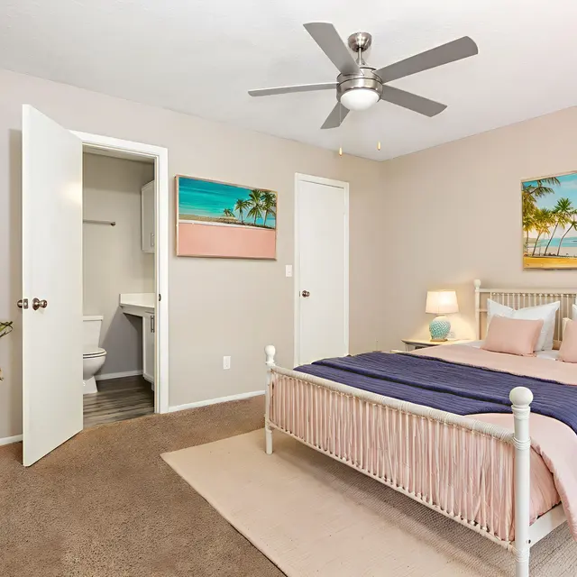 A modern bedroom featuring a white bed with pink and blue bedding. There is a ceiling fan, a potted plant, and two paintings of beach scenes on light-colored walls. A door leads to a bathroom.