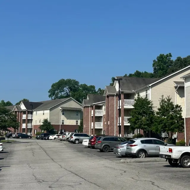 A view of an apartment complex with multiple buildings alongside a parking lot filled with parked cars, under a clear blue sky.