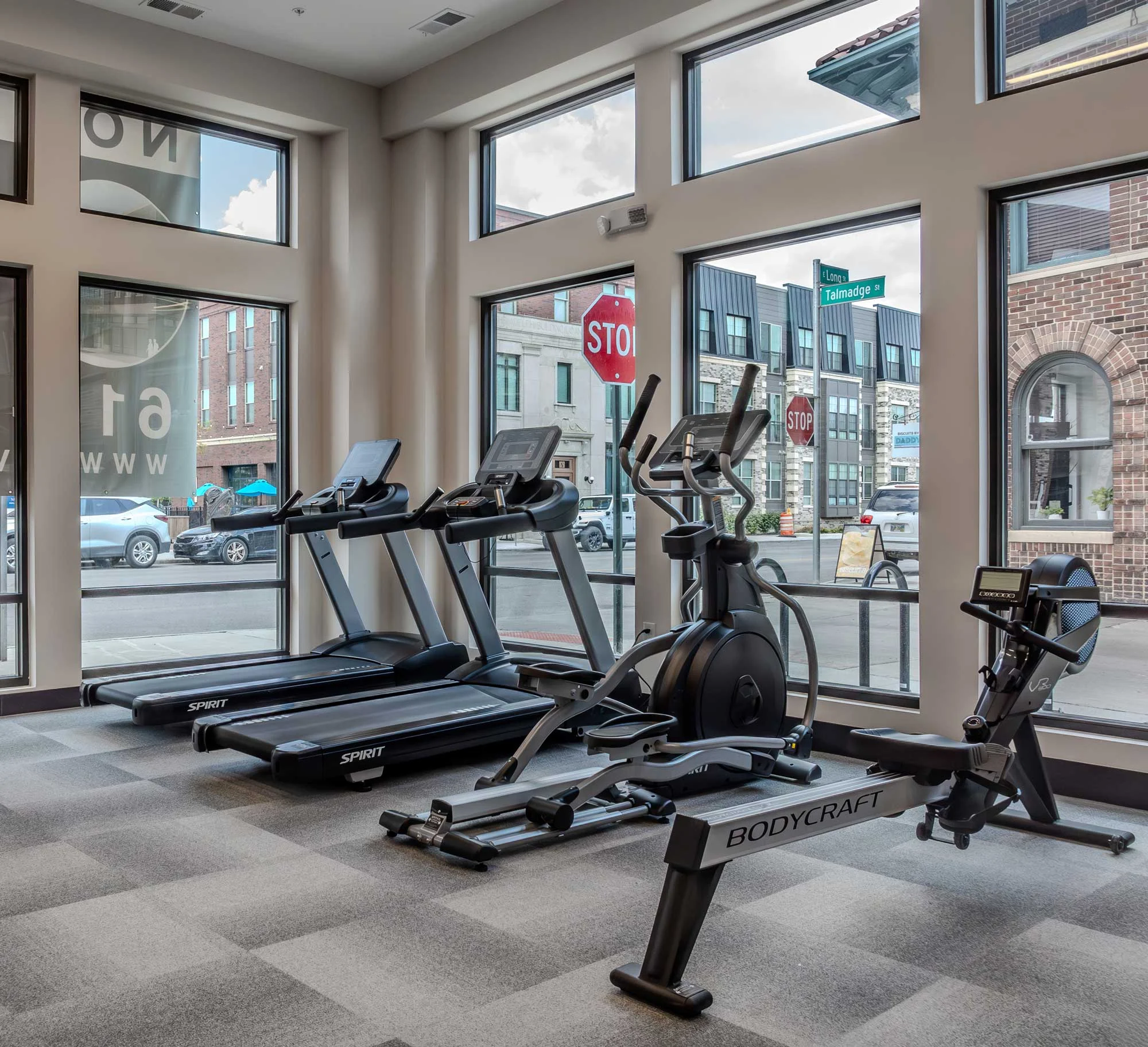 Modern Gym Interior Interior view of a fitness area featuring cardio machines including treadmills, an elliptical, and a rowing machine. Large windows showcase an outdoor street view.