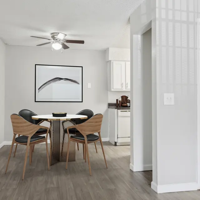 A modern dining area with a round table and four chairs, bright walls, a ceiling fan, and a kitchen visible in the background.