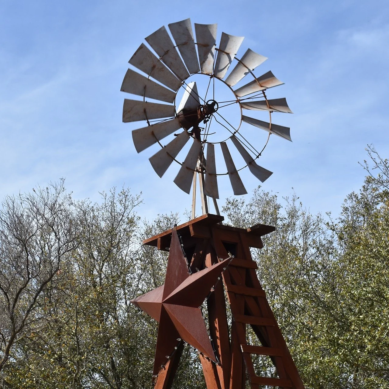 A vintage windmill with a rusty star-shaped base, surrounded by greenery and a clear blue sky.