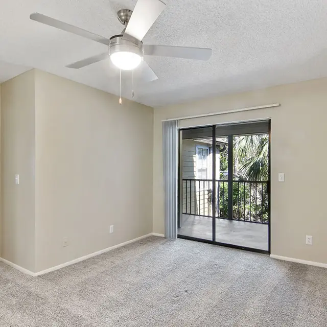 A beige room with sliding glass doors entering a balcony, a ceiling fan, and carpeted floors.
