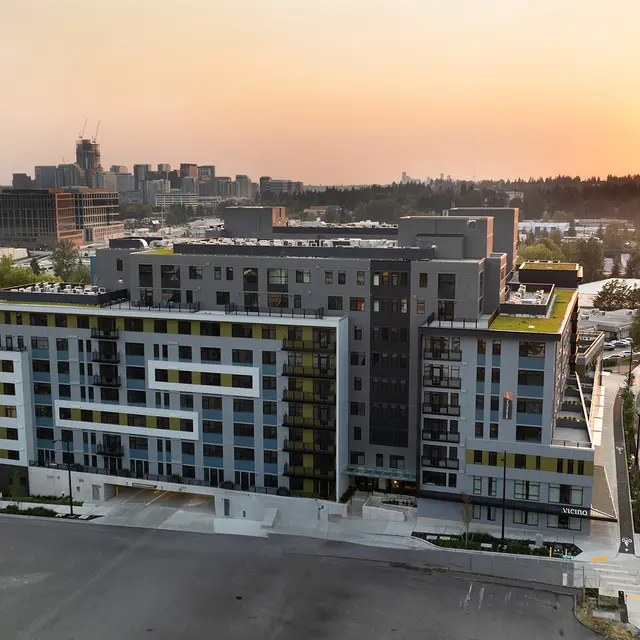 A modern multi-story apartment building with a sleek design seen at sunset. The building features balconies and a unique layout, surrounded by urban infrastructure and trees. The setting sun casts a warm glow over the scene.
