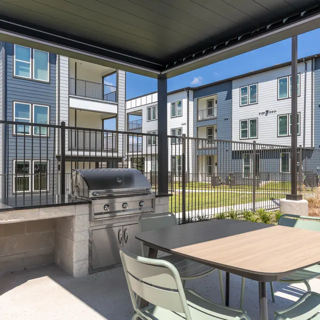 An outdoor grilling area with a barbecue grill and a table surrounded by chairs, overlooking an apartment complex.