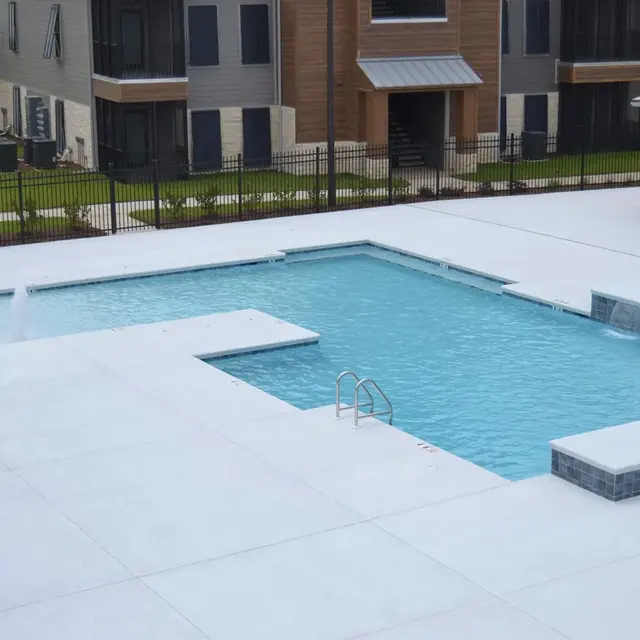A spacious swimming pool area at an apartment complex featuring a rectangular pool, a hot tub, and a decorative water feature, surrounded by concrete decking and green lawn.