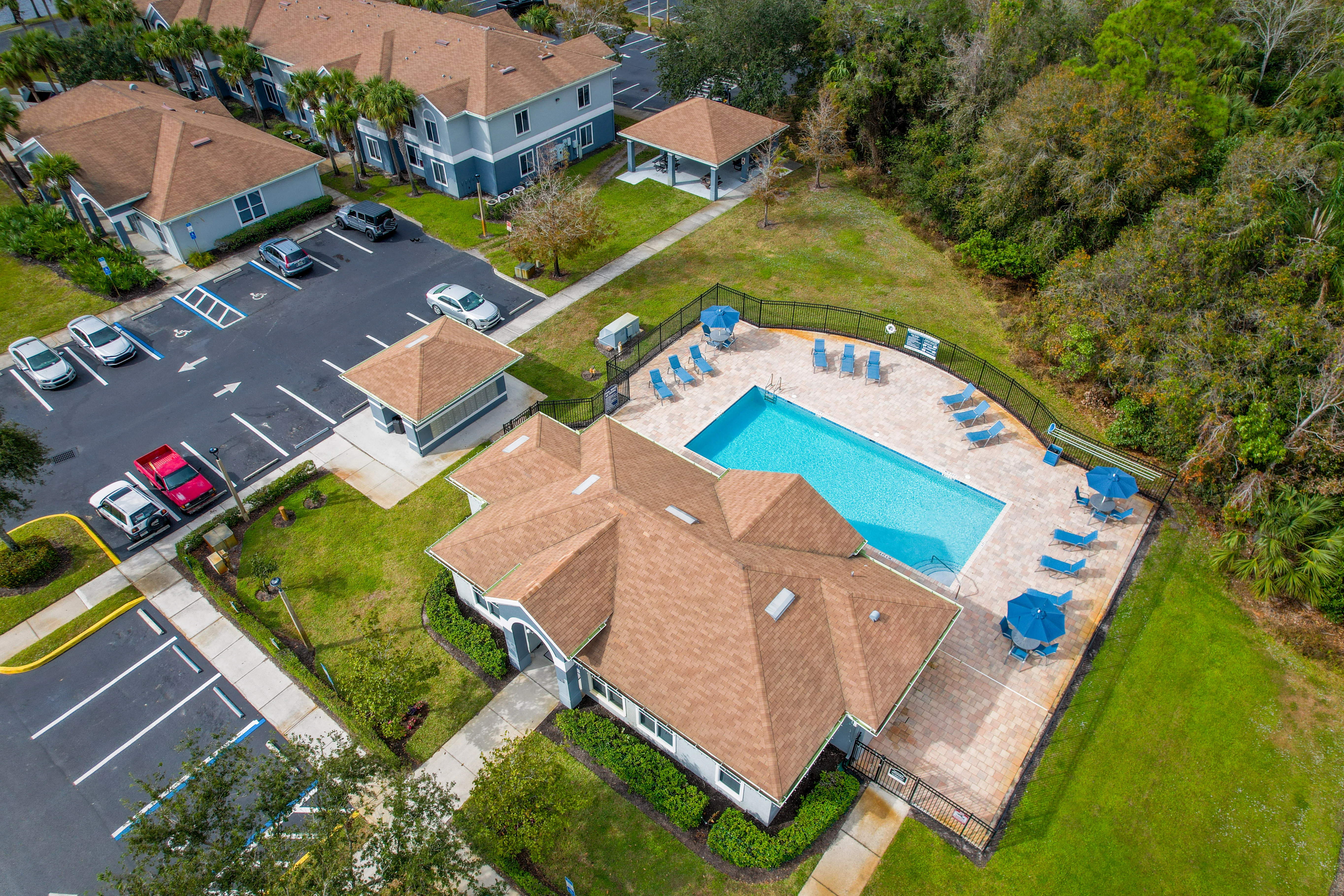 Aerial view of a community pool area with a rectangular pool surrounded by lounge chairs and grass. Nearby, there are parked cars and residential buildings with brown roofs.