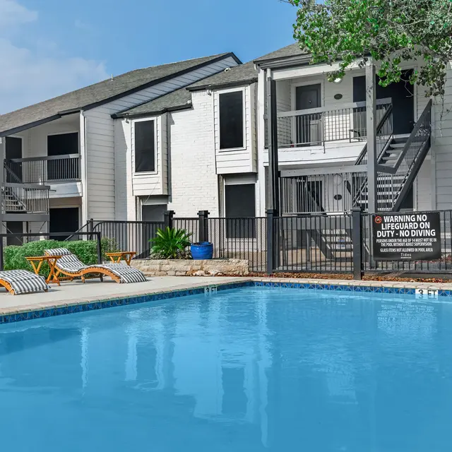 Fenced-in community pool with lounge chairs and small tables next to chairs. 