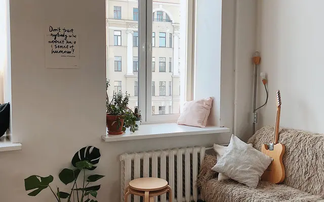 A cozy room with a sofa, plants, a round table, and a guitar, featuring large windows with natural light streaming in.