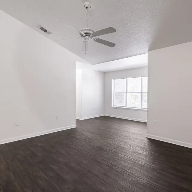 An empty room with dark wood flooring, white walls, and a ceiling fan. It has a large window allowing natural light to enter.
