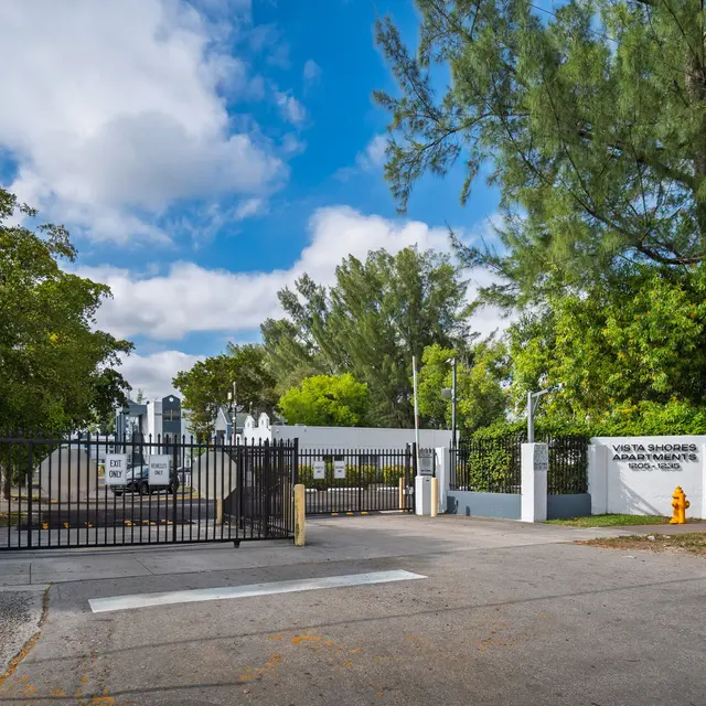 Entrance gate to a facility with a white wall and some greenery in the background under a partly cloudy sky.
