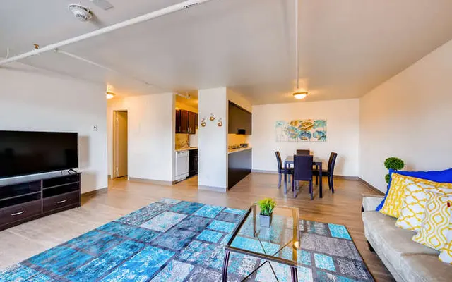 A spacious living area in a modern apartment, featuring a blue area rug, a TV stand with a television, and a dining table with black chairs. The kitchen area is partially visible in the background, with dark wooden cabinets and light countertops. Decor includes a colorful wall art piece and decorative cushions on the sofa.