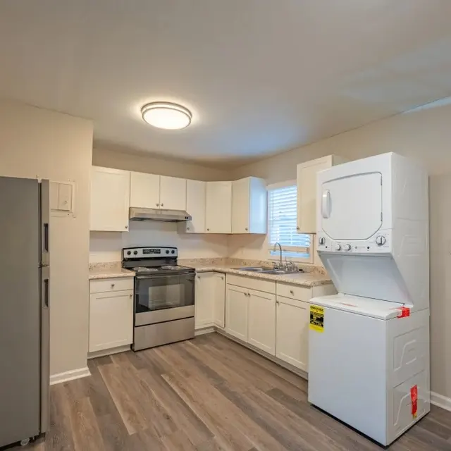 A modern kitchen featuring white cabinetry, a stainless steel refrigerator, a range with an oven, and a stacked washer and dryer. The kitchen has gray wooden flooring and a window allowing natural light.