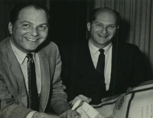 Two men smiling and posed for a photograph at a table with documents in front of them.