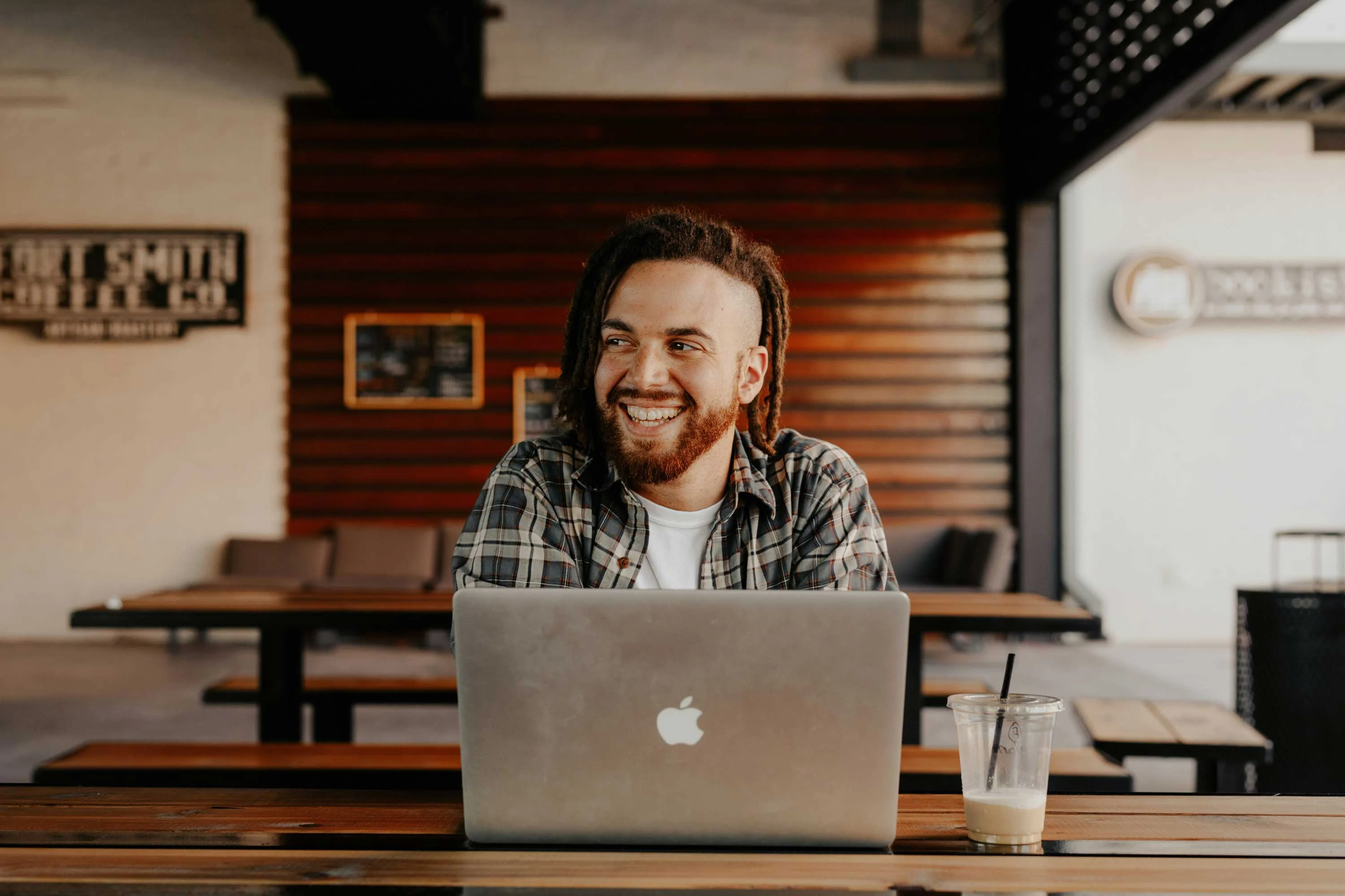 Smiling man with dreadlocks sitting at a table with a laptop and iced coffee in a bright, modern cafe setting.