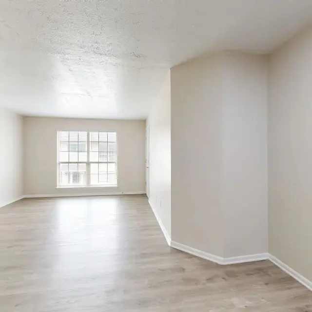 A spacious, empty living room with light-colored walls and a large window letting in natural light. The floor is made of light wood, and there are no furnishings or decorations.