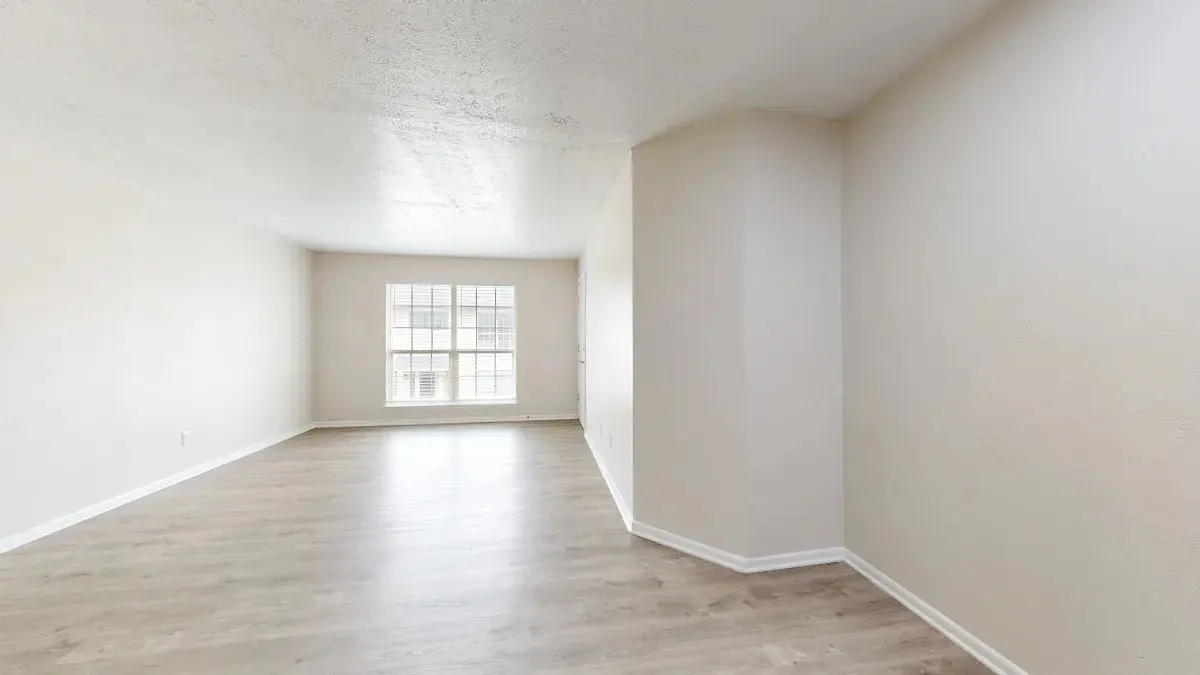 A spacious, empty living room with light-colored walls and a large window letting in natural light. The floor is made of light wood, and there are no furnishings or decorations.