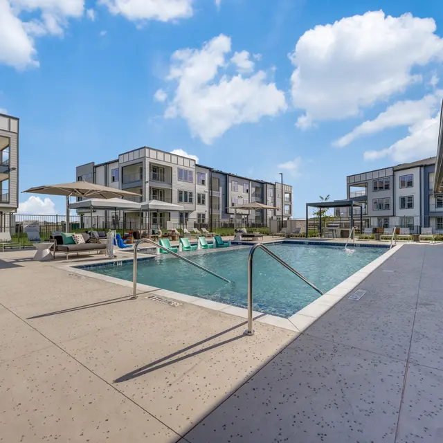 A modern apartment complex with a swimming pool and lounge chairs under shaded umbrellas.