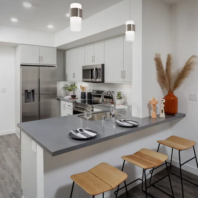 A modern kitchen featuring sleek white cabinets, stainless steel appliances, and a gray countertop. Three wooden stools sit at the bar area, which is adorned with table settings. A vase with decorative plants adds a touch of greenery to the countertop.