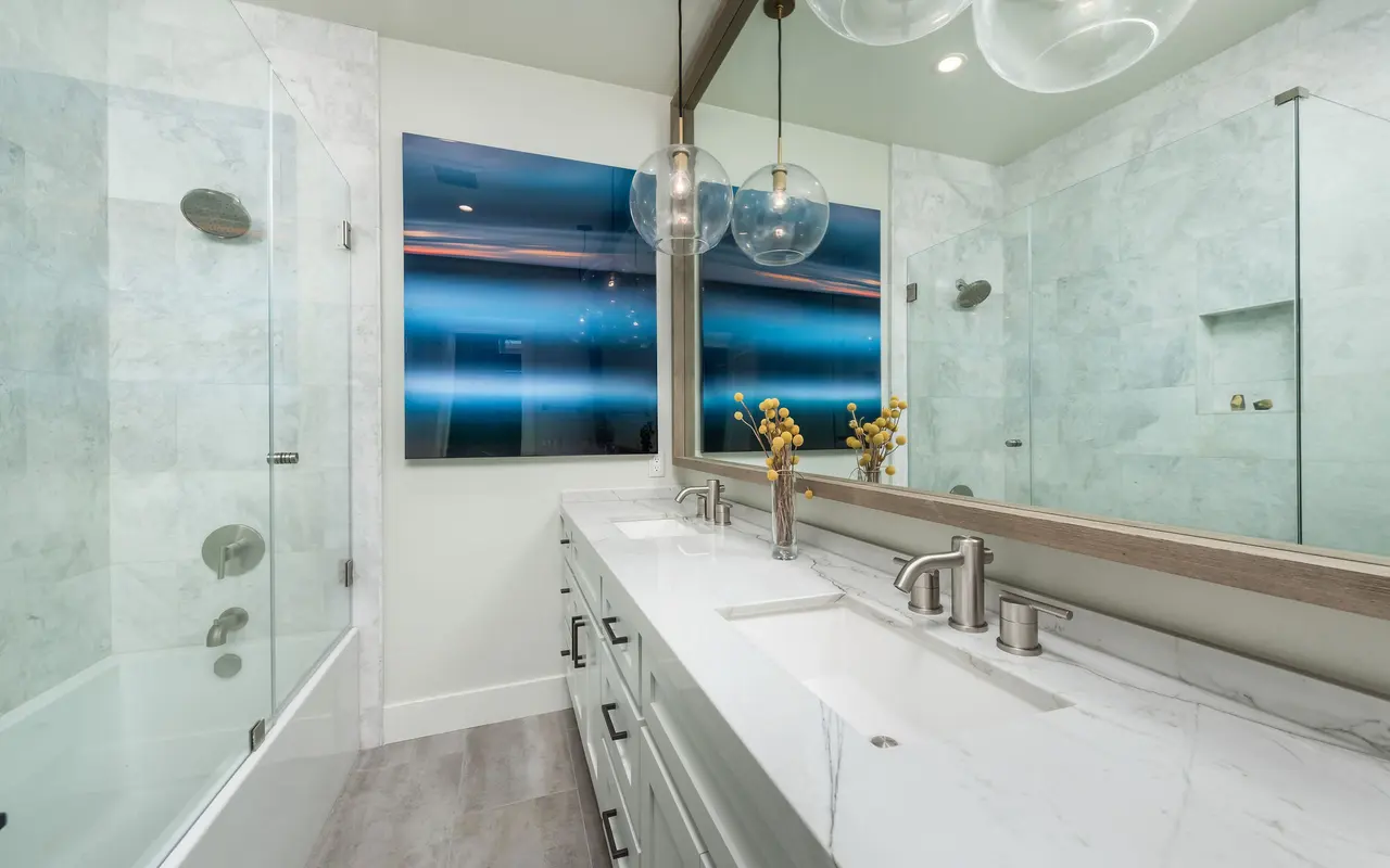 A modern bathroom featuring a large vanity with a marble countertop, two sinks, and elegant fixtures. The room includes a glass shower with a tub, and a wall-mounted TV displays digital art. Ambient lighting is provided by stylish pendant lamps.