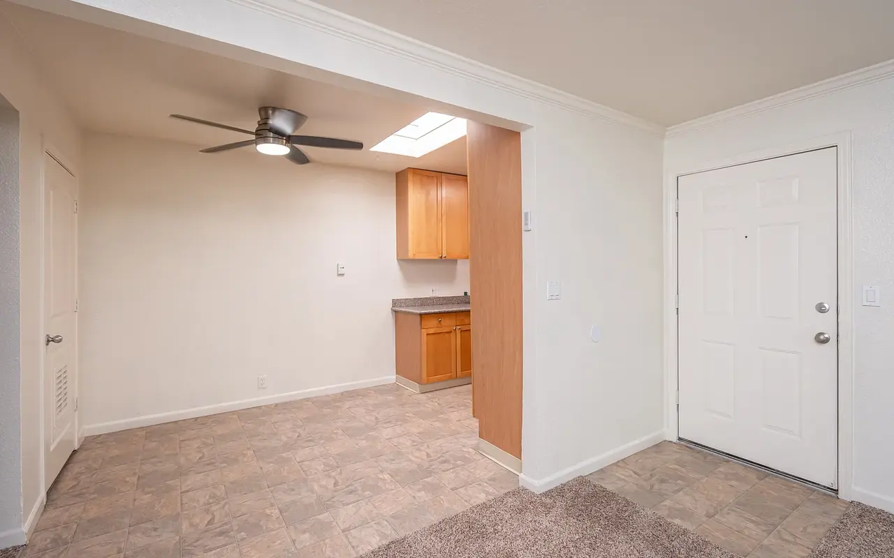 A view of a light-colored room with a fan, a small kitchen area on the left, and a door to the right.