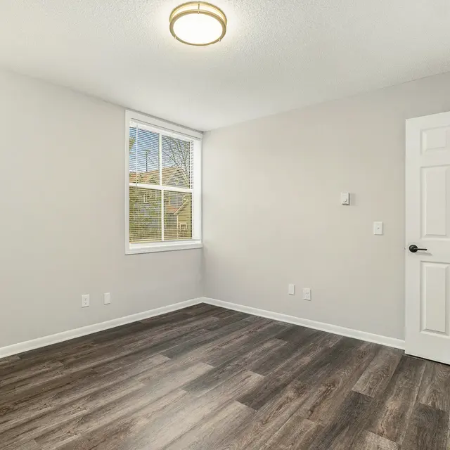 An empty room with light gray walls, a large window allowing natural light, and dark wooden flooring. A white door is visible on the right side of the image.