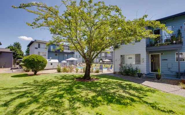 Apartment Complex with Tree and Pool View of an apartment complex featuring a large tree, lawns, and a pool area in the background.