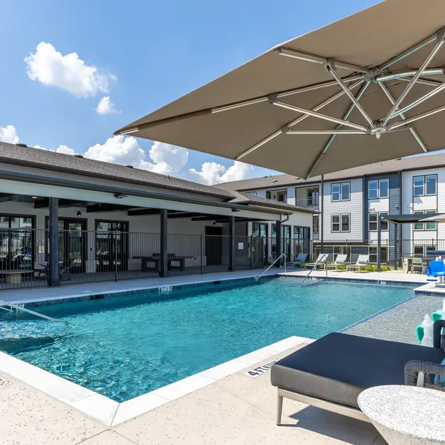 A modern apartment complex featuring a swimming pool surrounded by lounge chairs and umbrellas under a clear blue sky.