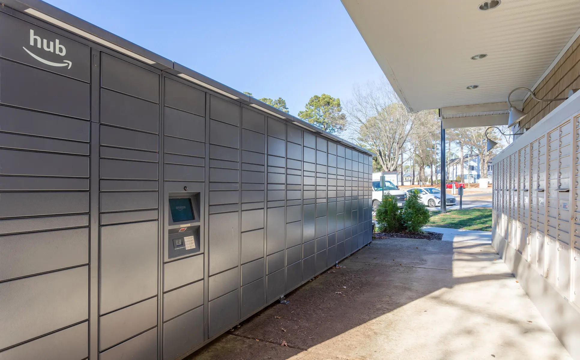 A row of Amazon hub lockers with a touchscreen interface located in a well-lit area beside a building. Trees are visible in the background, indicating an outdoor setting.