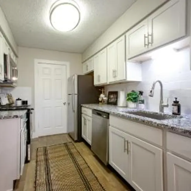 A modern kitchen featuring granite countertops, light-colored cabinets, stainless steel appliances, and a decorative rug on a wooden floor.