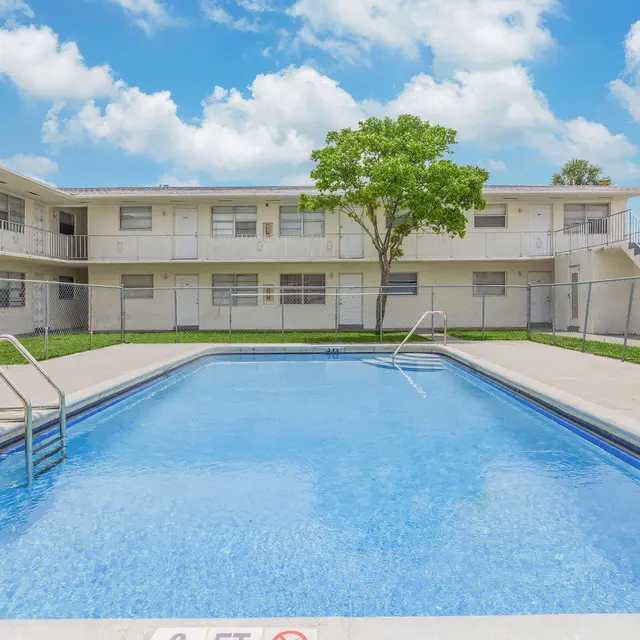 A clear swimming pool surrounded by a white two-story apartment building and green grass in a sunny setting.