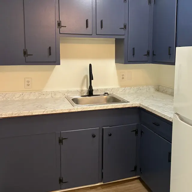 A small kitchen featuring blue cabinets and a white countertop. The sink is centrally located with a black faucet, and there are wooden floors visible.
