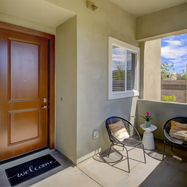 Town Germann - Balcony A cozy porch featuring a brown door, welcome mat, two chairs, a small table, and a potted plant under a clear blue sky.