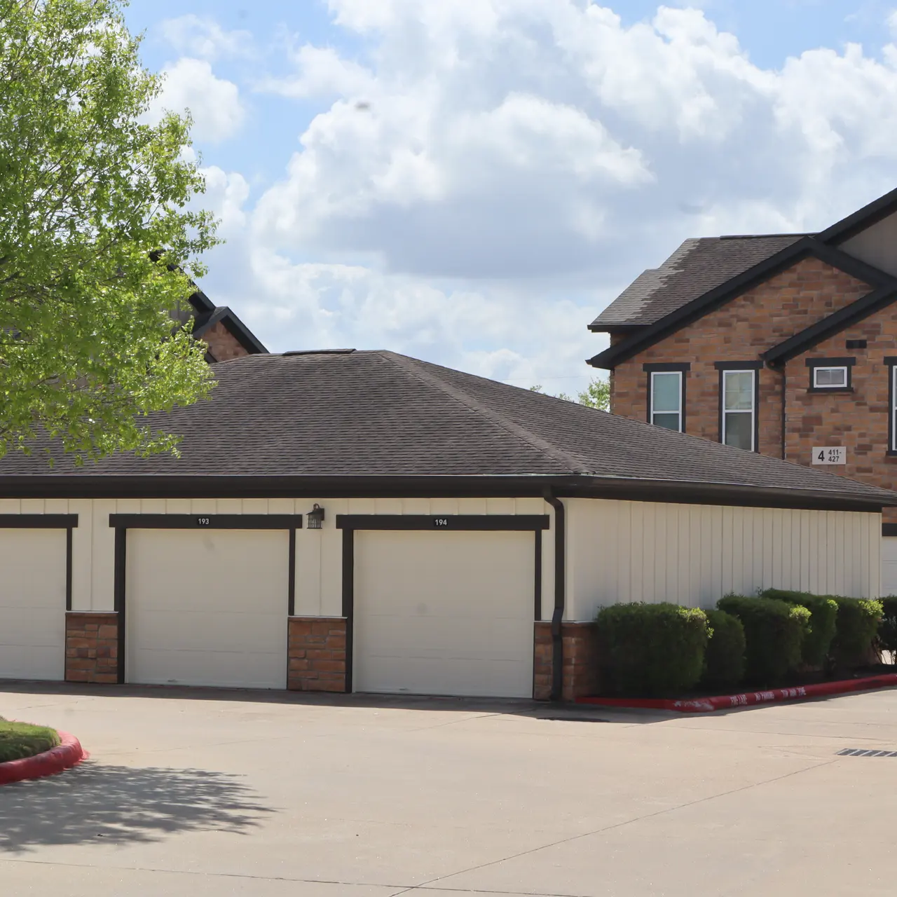An apartment complex with a row of garages in the foreground and a multi-story building in the background. There is a large tree on the left side and blue sky with clouds above.