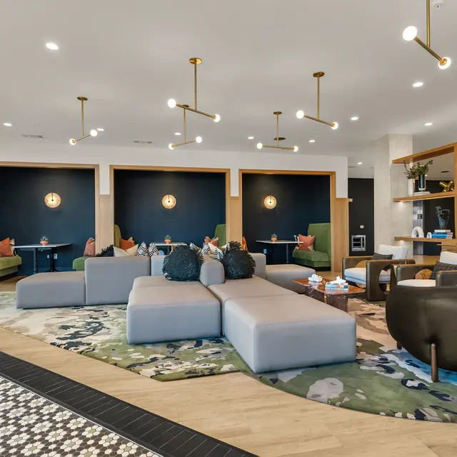 A modern lobby interior featuring a spacious seating area with a large gray sectional sofa and decorative cushions. The walls are dark blue with pendant lights overhead. There are small tables for drinks in front of the sofa, and a rug with a colorful design on the floor. A bookshelf with decorative items is visible in the background.